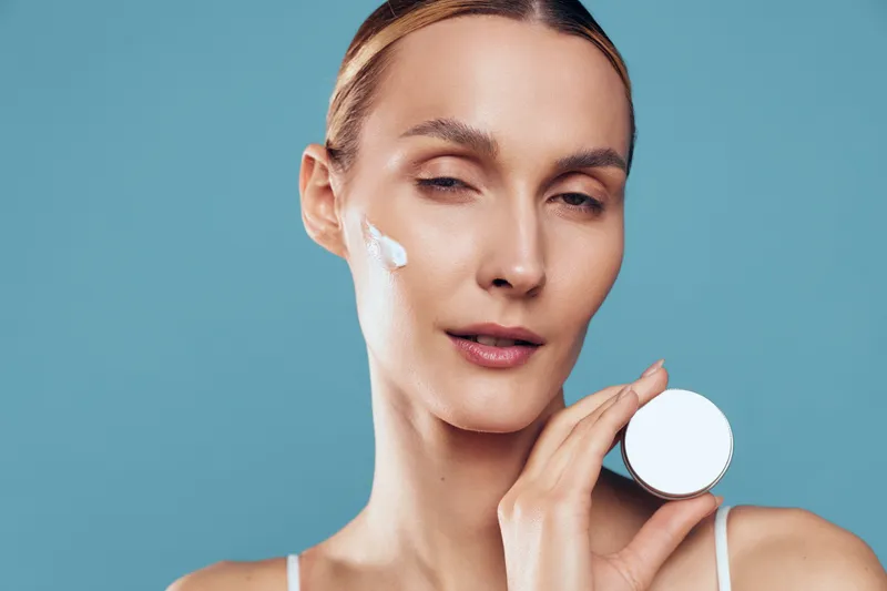 Woman applying cream on face with closed eyes holding jar against blue background, skincare routine, natural beauty, smooth skin, gentle expression, minimal makeup.