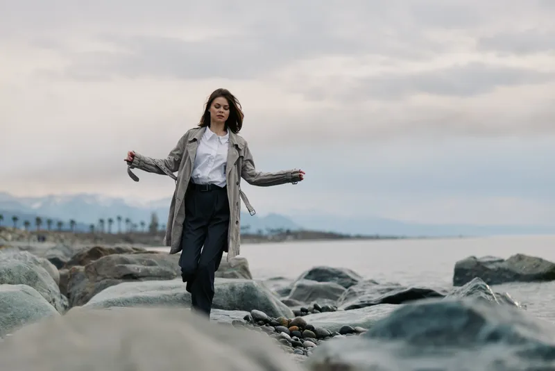 Young woman in trench coat walks on rocky beach with calm sea and cloudy sky. Casual fashion, outdoor lifestyle, and nature exploration concept with relaxed mood and cool weather.