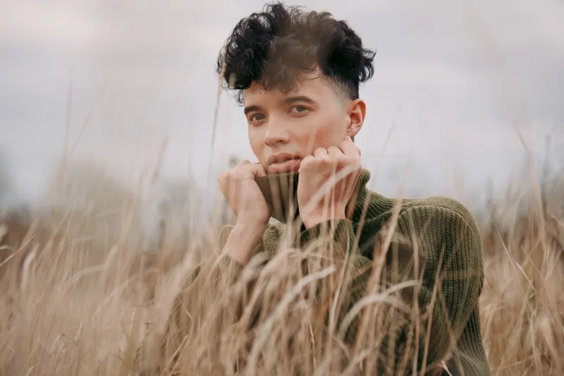 Close portrait of a young man in a wool sweater among tall field grasses, conveying quiet confidence and natural authenticity through a candid gaze and understated emotional credibility.