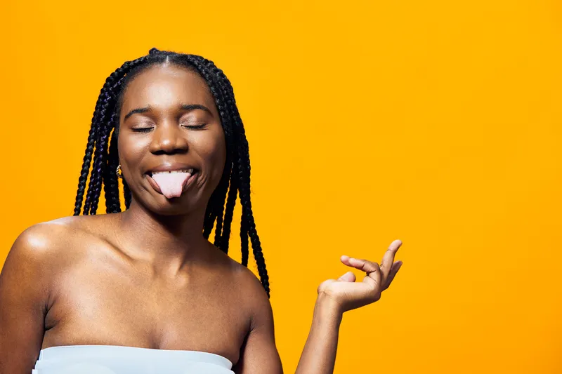 Joyful Black woman with braided hair playfully sticking out her tongue against a vibrant yellow background, showcasing confidence and fun spirit