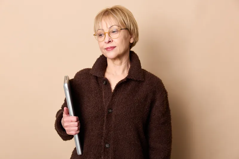 Elderly woman with glasses holding a clipboard and book, studio portrait, calm expression on beige background, education and learning concept, copy space