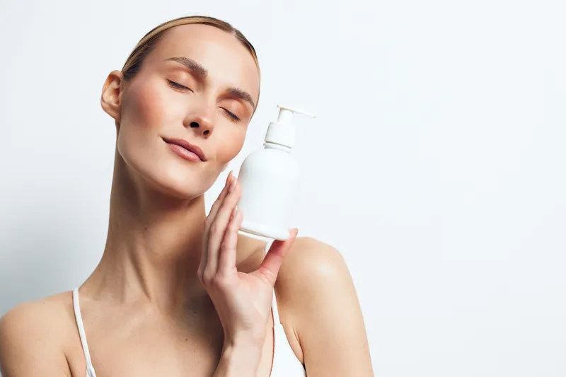 Young woman with clear skin holding white cosmetic bottle near face, eyes closed, gentle smile, soft lighting, minimalistic background, skincare concept.