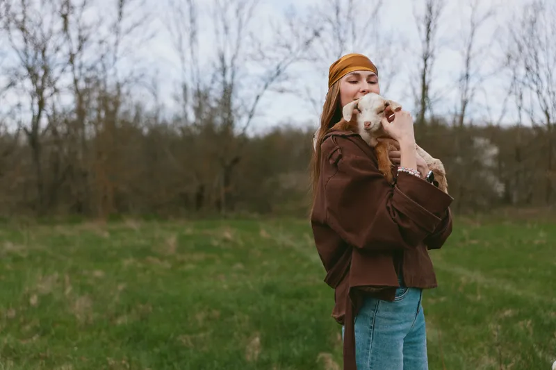 Caring woman outdoors hugs a goat kid in a grassy field, wearing a brown coat and beanie. Natural light emphasizes a warm, wholesome lifestyle moment with animals and nature.