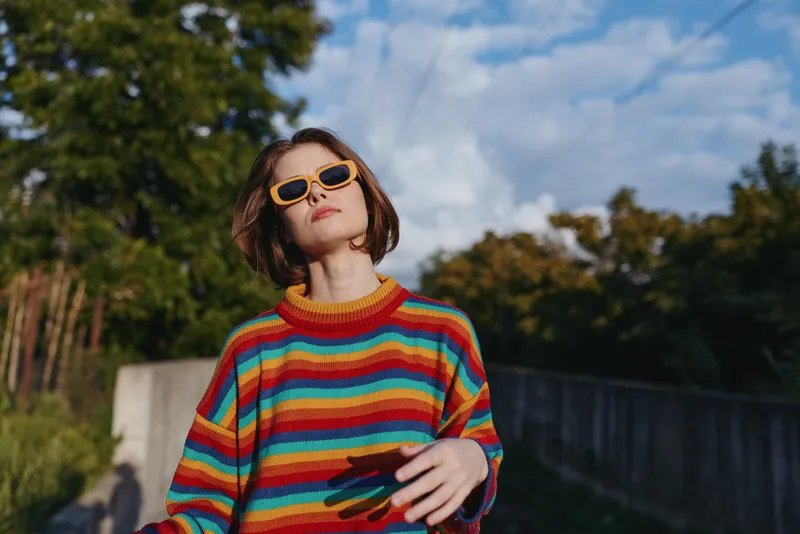 Young woman in a rainbow striped sweater and yellow sunglasses, bob haircut, standing outdoor in an urban park setting, confident pose, casual outfit for lifestyle and fashion travel.