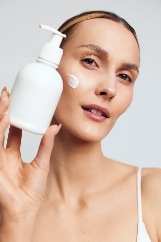 A young woman with clear skin holds a white lotion bottle near her face while applying cream on her cheek. The bright background emphasizes skincare and beauty routine concepts.