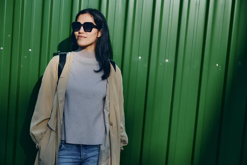 Woman wearing a beige coat over a gray sweater and jeans, sunglasses on, stands against a tall green wall, exuding casual street style and confident urban vibe.