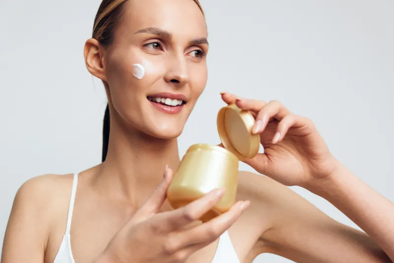 Smiling woman applying cream on face holding open jar skincare beauty routine natural makeup wellness clean skin white background