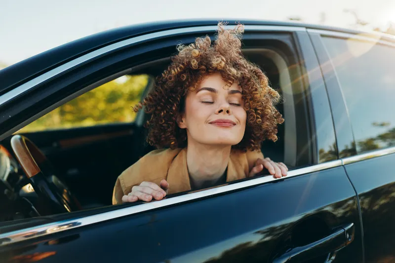 Young woman with curly hair leans from a sunlit car window, eyes closed and smiling, enjoying a gentle breeze outdoors in a casual pose