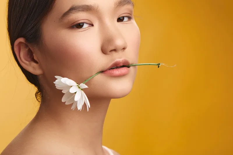 asian woman with a daisy held between lips stands against a warm yellow backdrop, presenting a calm, elegant beauty and soft, natural charm in a striking closeup.