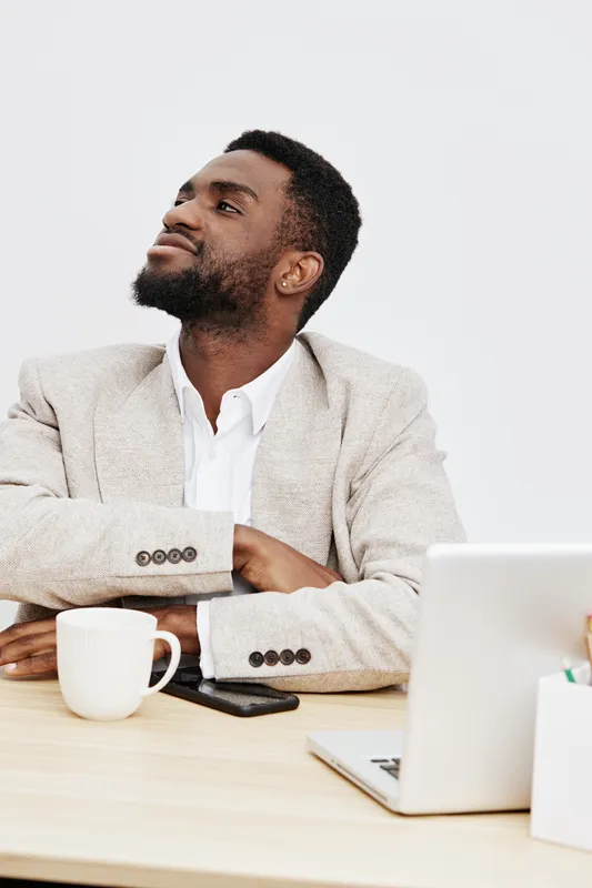 Confident man in beige suit sitting at modern office desk with laptop, coffee mug, and smartphone, looking thoughtfully away against plain white background, professional workspace, business concept.