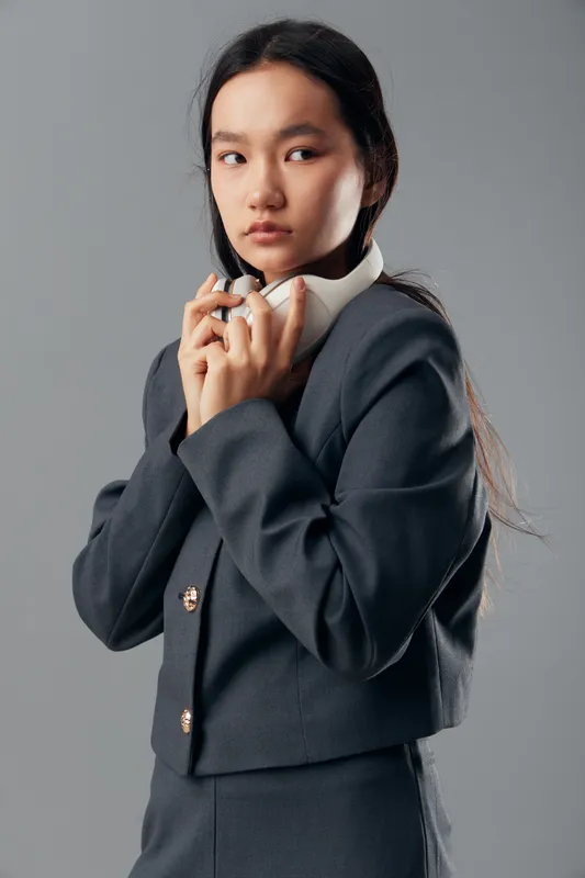 Young woman wearing dark business suit holding white headphones looks to the side with serious expression against gray background in studio. Female model with long hair in portrait.