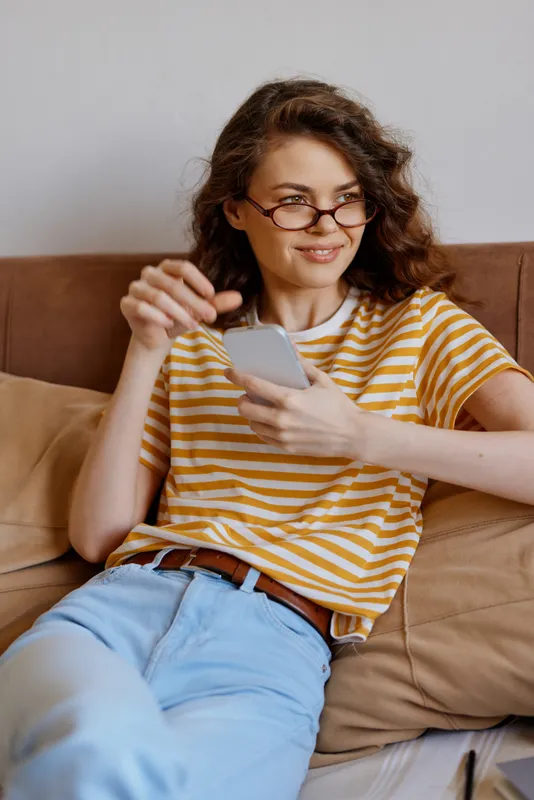 Young woman at home relaxing on a sofa, using a smartphone, wearing striped t shirt and glasses, casual lifestyle scene with natural light and cozy decor. Soft background music implied.