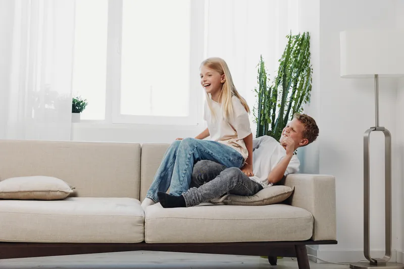 Happy children playing together on beige sofa in bright living room. Boy and girl laughing while having fun, casual clothing, natural light and cozy home atmosphere.