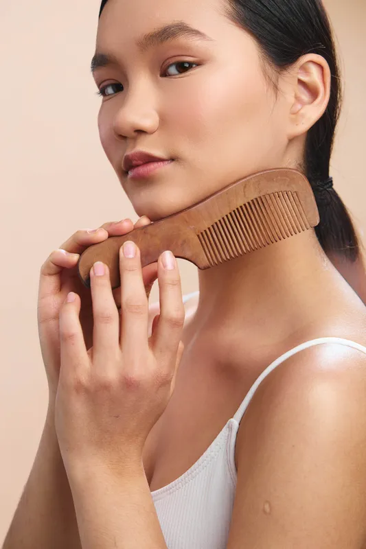 Asian woman gently styles hair with a wooden comb in a soft studio setting. The calm portrait highlights elegance, poise, and self care, capturing a focused moment of beauty and personal grooming.