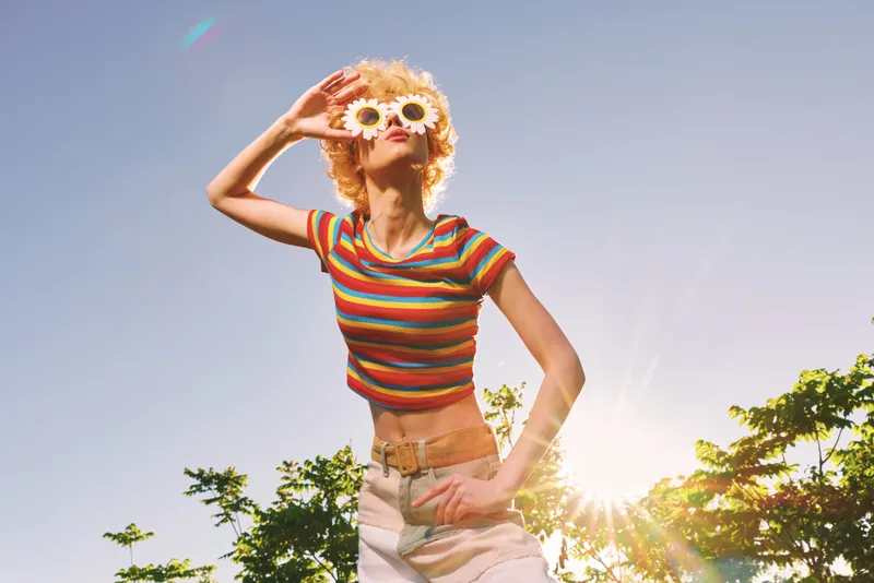 Playful woman in a striped crop top wearing flower sunglasses outdoors, sun flare and blue sky, vibrant retro lifestyle, carefree mood, natural light, sunny day