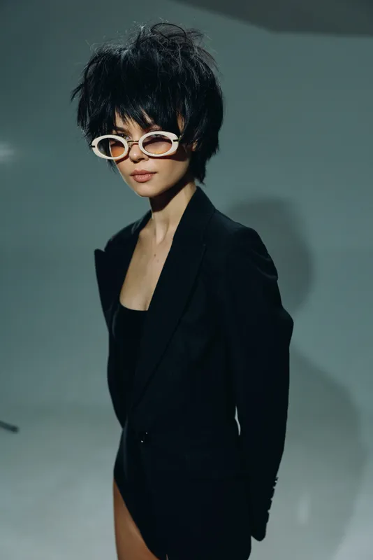 Stylish woman with short black hair wearing white sunglasses and a black blazer, posing confidently in a minimalistic studio setting.