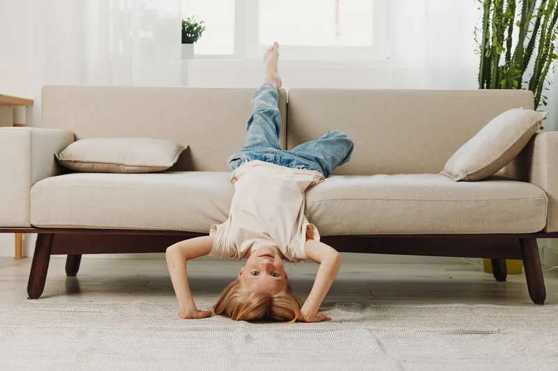 Playful child in casual clothes lies upside down on a beige sofa in bright living room with natural light. Concept for childhood joy, home comfort, and carefree moments indoors.