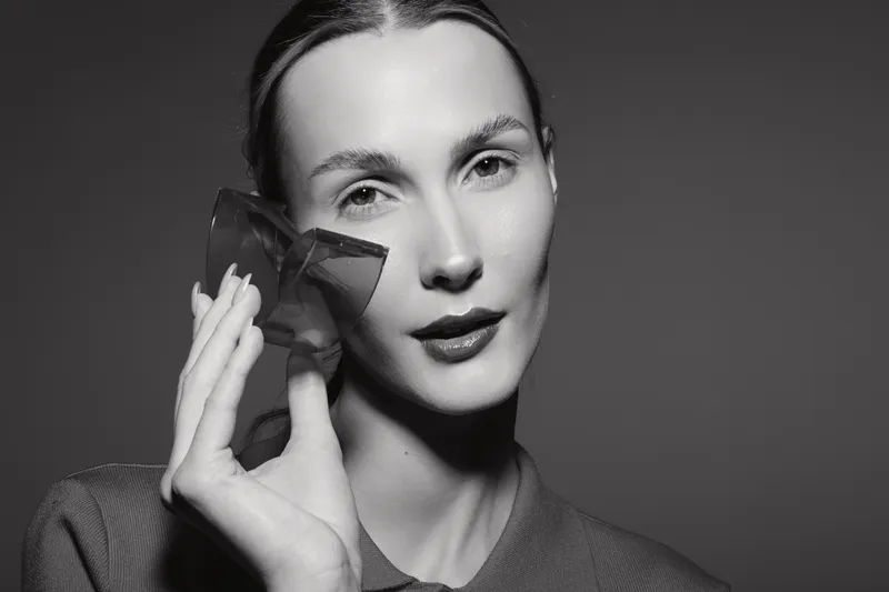 Portrait of a pale white woman holding a transparent geometric glass object near her face in studio lighting.