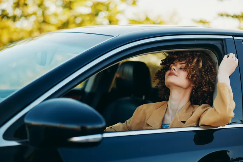 Curly haired woman in a tan coat relaxing in a car, sunlight filters through trees, a lifestyle moment conveying freedom, travel, and calm with retro film color.