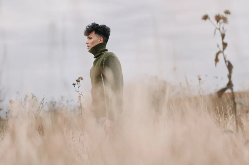 A contemplative young person in a textured green sweater walks through tall dry grass, captured with natural light and an honest perspective, emphasizing emotional authenticity and trustworthiness.