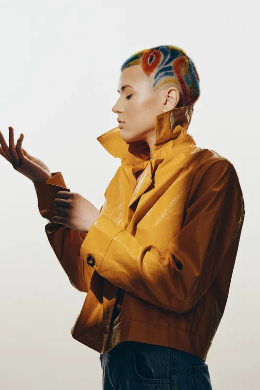 woman with colorful shaved hair wearing a mustard yellow jacket, posing thoughtfully against a simple background, showcasing modern fashion and unique style.