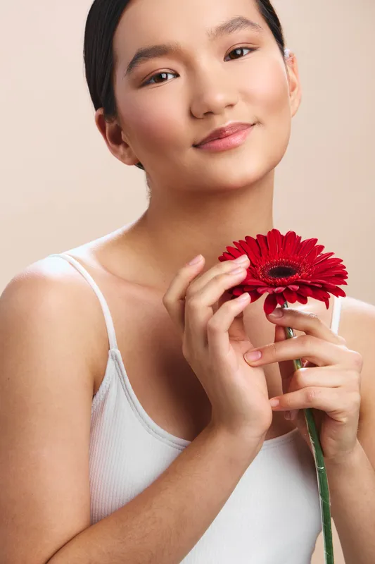 asian woman holds a vibrant red flower close to her face in soft studio light, exuding calm grace and natural beauty, a gentle smile enhancing approachable warmth and elegance.