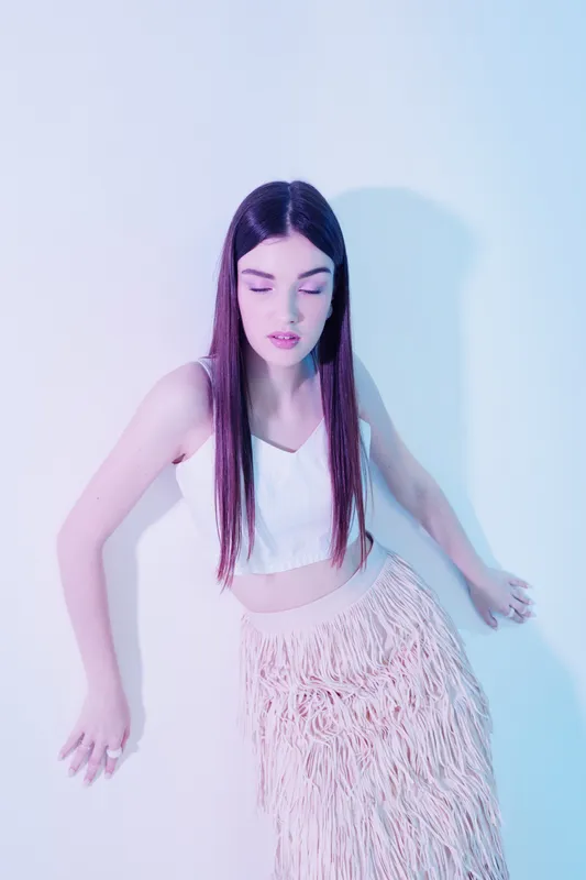 Young woman with long hair in white top and fringed skirt poses with closed eyes against pastel background. Concept of beauty fashion and elegance in studio lighting.