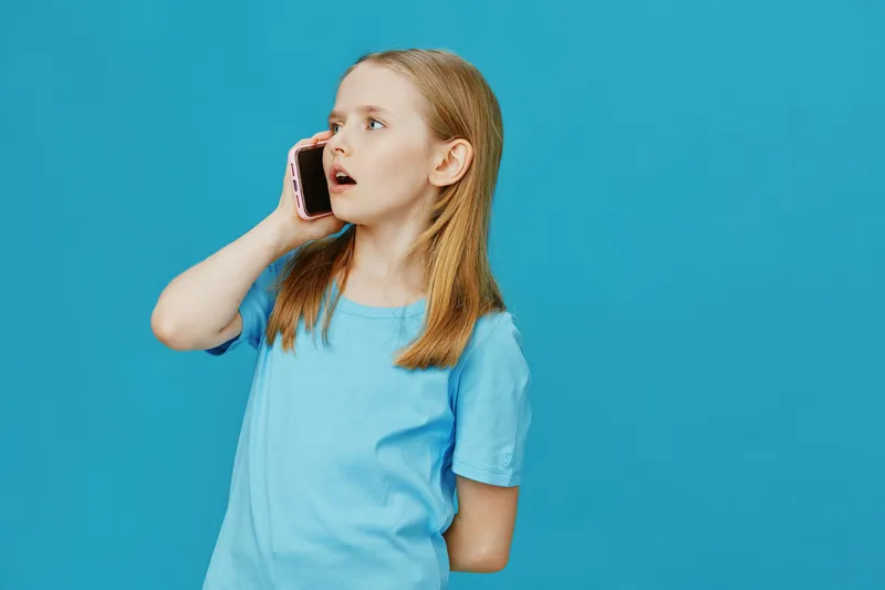 A young girl with light brown hair in a blue t shirt talking on a smartphone, expressing surprise against a bright blue background, ideal for concepts of communication and youth