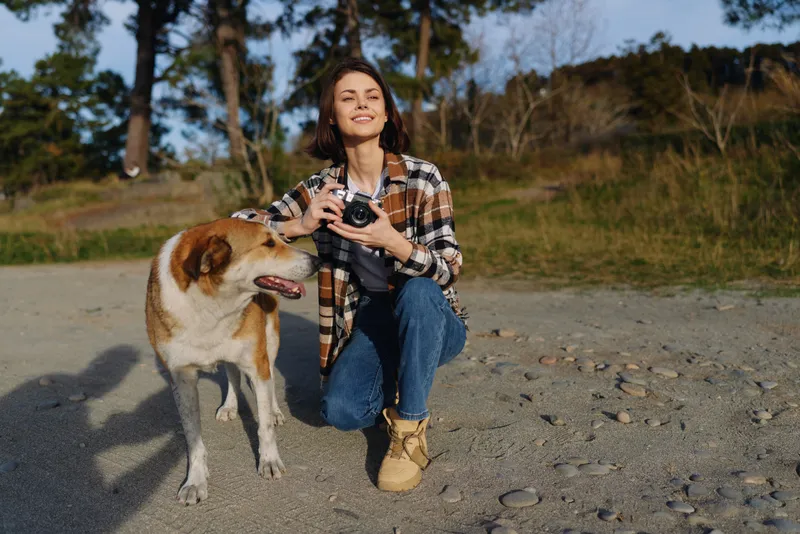 Woman photography with camera kneels beside a dog on a gravel path, outdoors in nature. A candid portrait captures companionship, adventure, and joyful exploration under sunny trees.