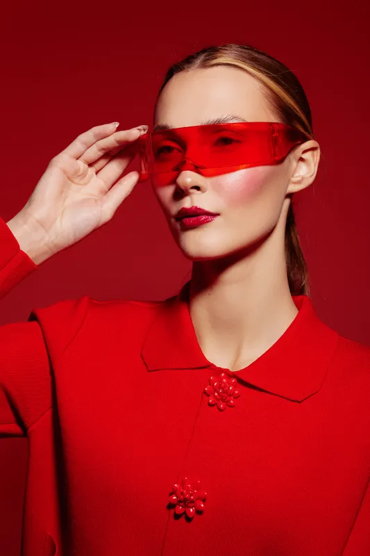 Stylish woman in red outfit and translucent red visor poses against red background. Fashion portrait with glossy makeup and sleek hairstyle. Concept of modern style and bold color.