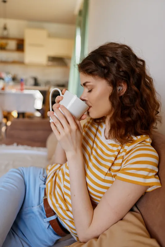 Young woman with curly hair sits on a cozy sofa at home, sipping from a mug and enjoying a calm, sunlit morning in a bright living room with soft cushions and warm wood tones, creating an intimate
