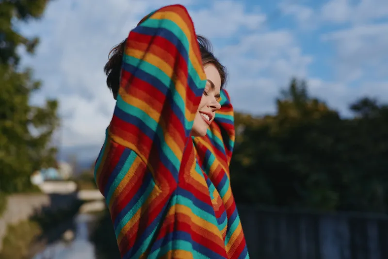Woman wearing a colorful rainbow sweater smiling outdoors, young woman with arms up in casual outfit, relaxed urban park setting, joyful expression for lifestyle, fashion and travel.