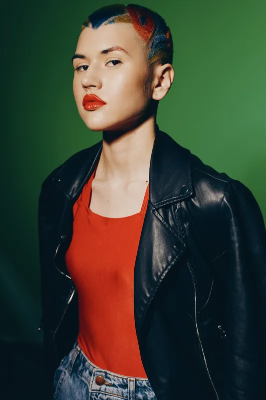 Young woman with colorful short hair wearing a black leather jacket and red top, posing confidently against a solid green background in vibrant lighting.