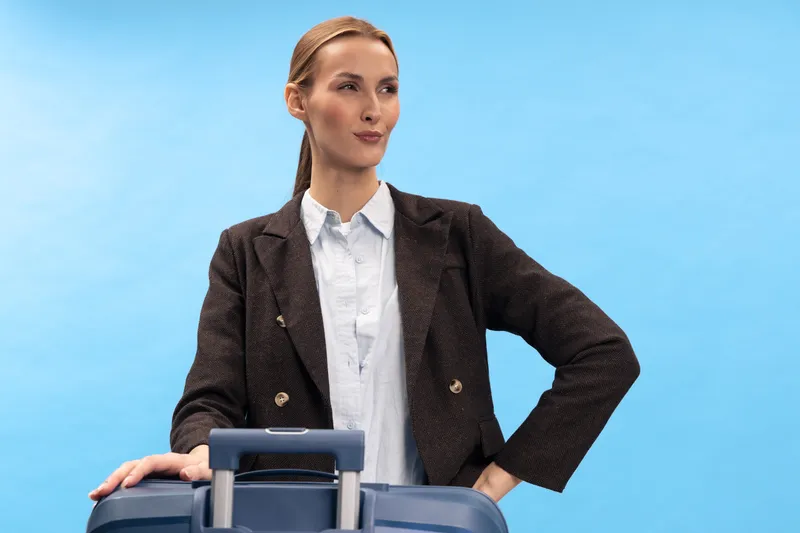 Confident businesswoman in dark blazer stands with suitcase on blue background. Professional woman poses with hand on hip and looks away. Concept for travel, business trip, and confidence.
