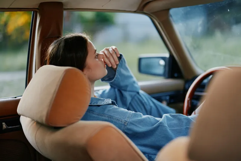 Young woman resting in a vintage car, wearing a denim jacket, looking thoughtful, with a serene expression and natural background, evoking nostalgia and calmness.