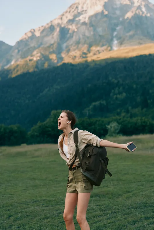 A young woman in hiking attire cheers with arms outstretched in a grassy valley against a mountain backdrop, with a backpack and phone in hand, capturing a moment of freedom and adventure.