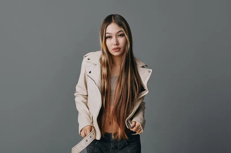 Young woman with long hair and beige jacket standing against gray background. Fashion portrait with casual clothing, model posing for studio photo shoot with serious expression.
