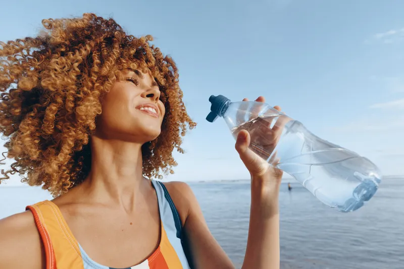 A young african american woman with curly hair drinks water from a bottle on a sunny beach, wearing a bright athletic top, enjoying hydration during outdoor workout by the sea.