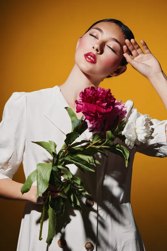 Elegant woman with glossy makeup holds bright peony flowers close to her white blouse. Her eyes are closed, posing softly against a golden background. Concept of beauty art fashion.
