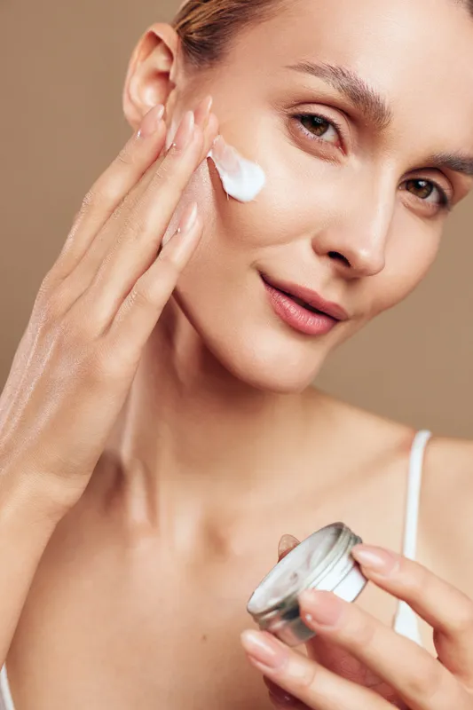 Young woman with brunette hair applies white face cream on her cheek while holding a small jar in a skincare routine, showing a calm and confident expression in a close-up portrait.