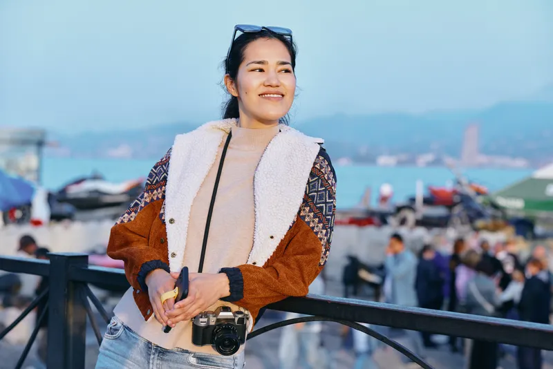 Woman with camera wearing a cozy jacket on outdoor coastal boardwalk, sunny day, smile, lifestyle scene, travel mood, fashion style, light breeze, urban scenery, candid moment