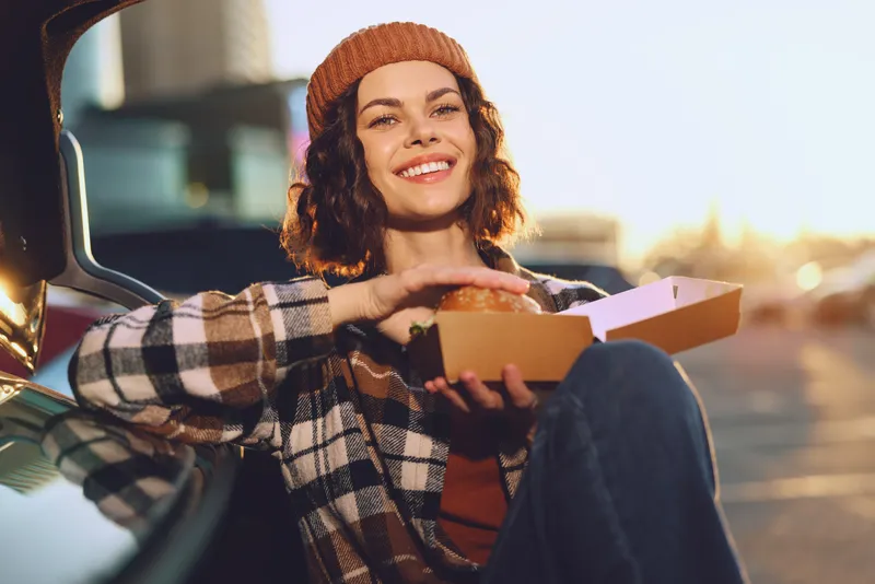 woman smile burger takeaway car candid moment lit by golden hour glow, authenticity and candid lifestyle captured as she eats outdoors in relaxed urban scene, mindful living and emotional storytelling