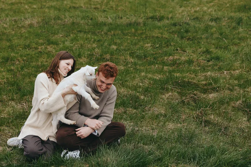Happy couple sits on the grass in cozy sweaters, embracing a kitten, smiling in a natural outdoor moment, a warm lifestyle scene on a sunny park setting.