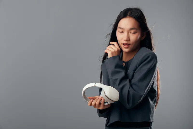 Woman holding headphones with closed eyes and relaxed expression on gray background. Casual outfit, long hair and technology concept for music listening or concentration.