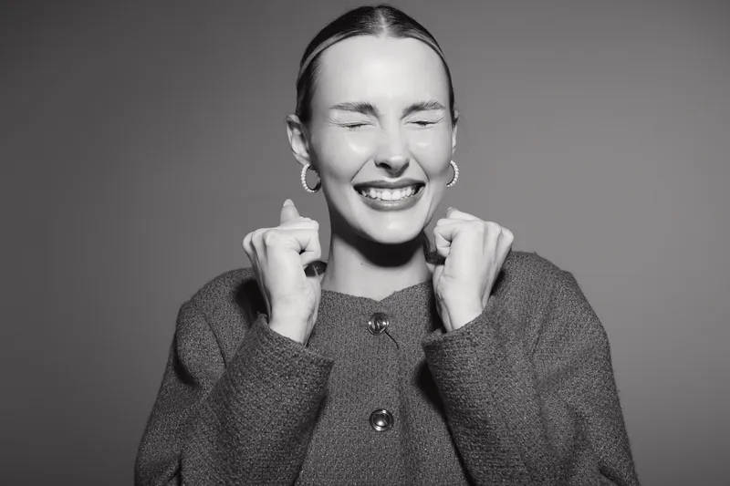 Happy Caucasian woman with light skin clenches fists and smiles with closed eyes in studio. Concept of joy, excitement, success, and positive emotions.