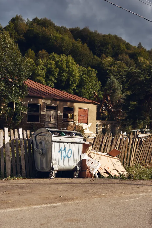 Dumpster and scattered debris beside a weathered house along a rural road, with a wooden fence and overgrown trees creating a neglected, gritty roadside scene. Dusty air hazy.