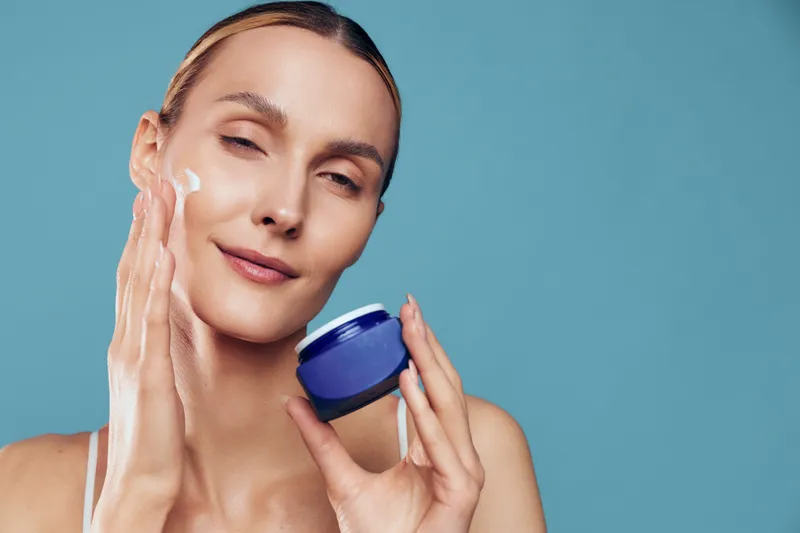 Skincare woman applies cream to face from a jar, smiling in a closeup. Blue studio background shows wellness and self care, with soft light and smooth skin.
