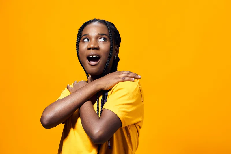 Excited young girl with braided hair in a yellow t shirt expressing joy against a vibrant orange background, showcasing emotions of happiness and surprise