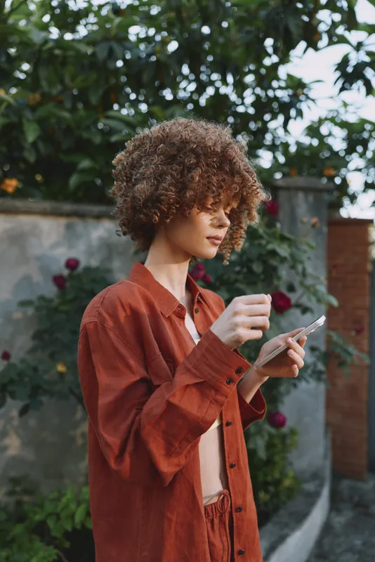 Woman with curly hair checks smartphone outdoors, embracing a casual lifestyle, wearing a rust color shirt, standing by a garden wall, capturing a candid everyday moment