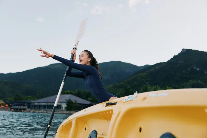 Dynamic action shot of a person paddling a yellow kayak on calm water with distant mountains, capturing energy, balance, and outdoor adventure in bright sunlight.